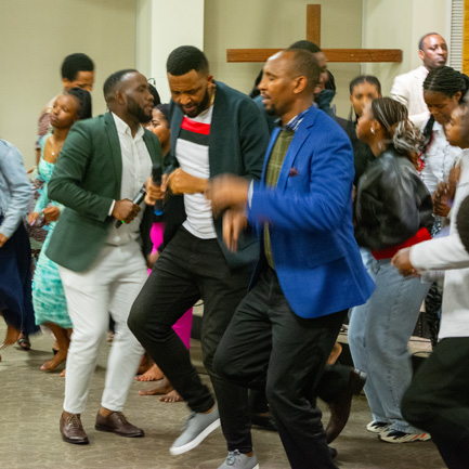 A large group gathered together in the church, dressed in their finest. They are seated, raising their hands praising God.