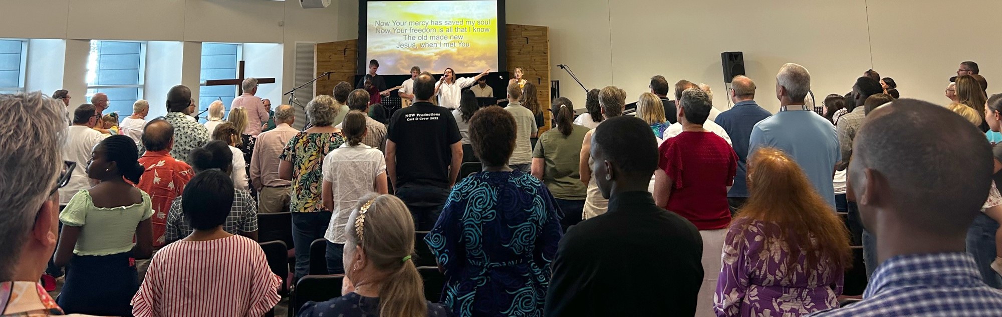 A large group of people of all ages, fill the church auditorium facing towards the stage where the worship team leads them in songs of praise.