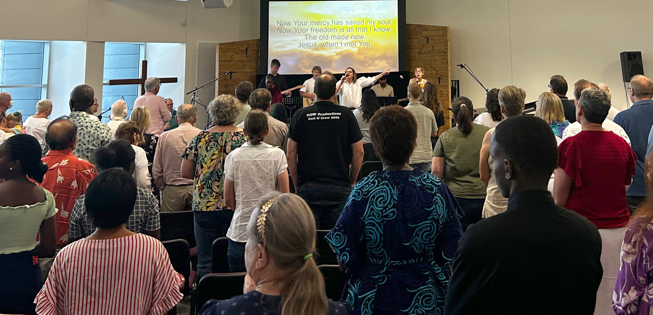 A large group of people of all ages, fill the church auditorium facing towards the stage where the worship team leads them in songs of praise.