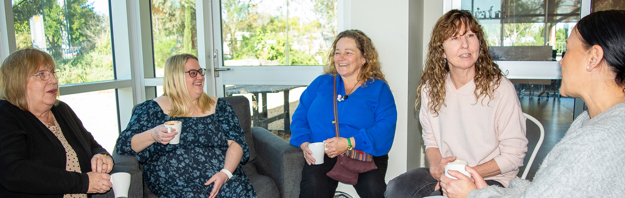 A group of 5 women sit together, holding cups of coffee. They are smiling and talking with each other.
