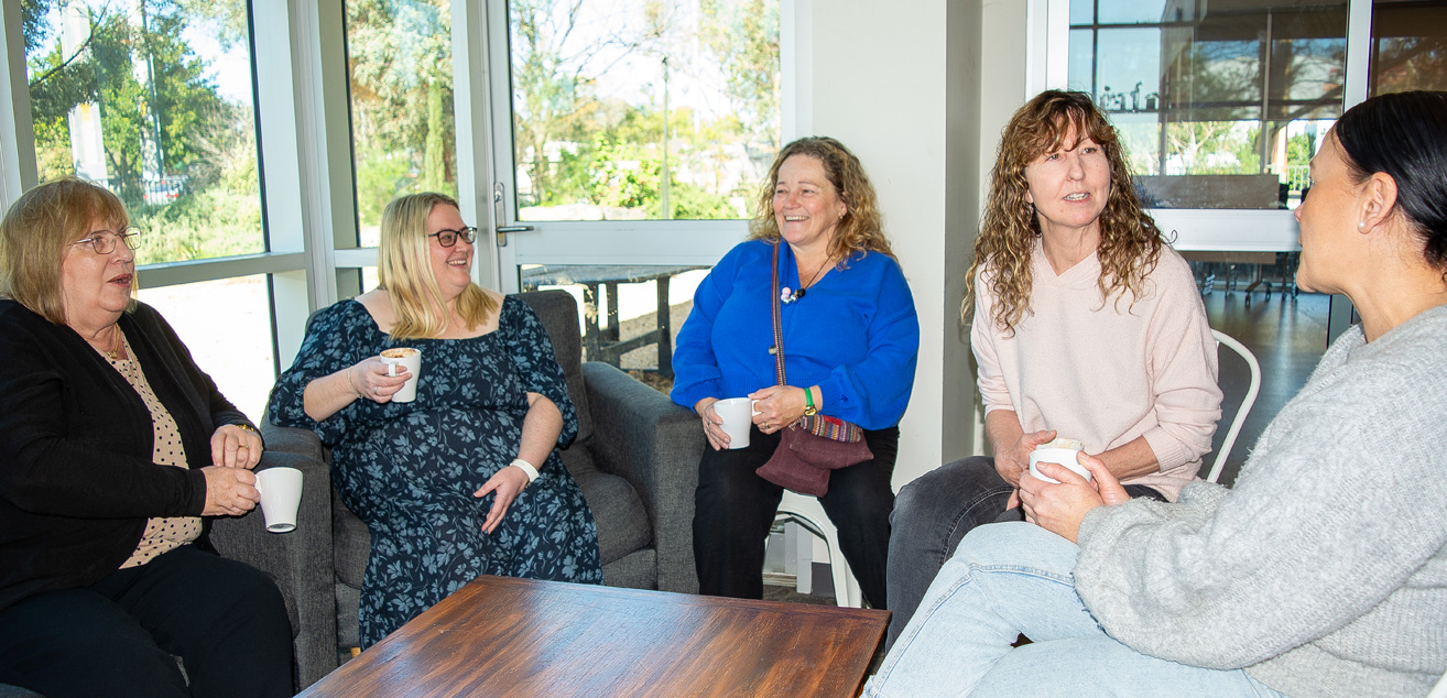 A group of 5 women sit together, holding cups of coffee. They are smiling and talking with each other.