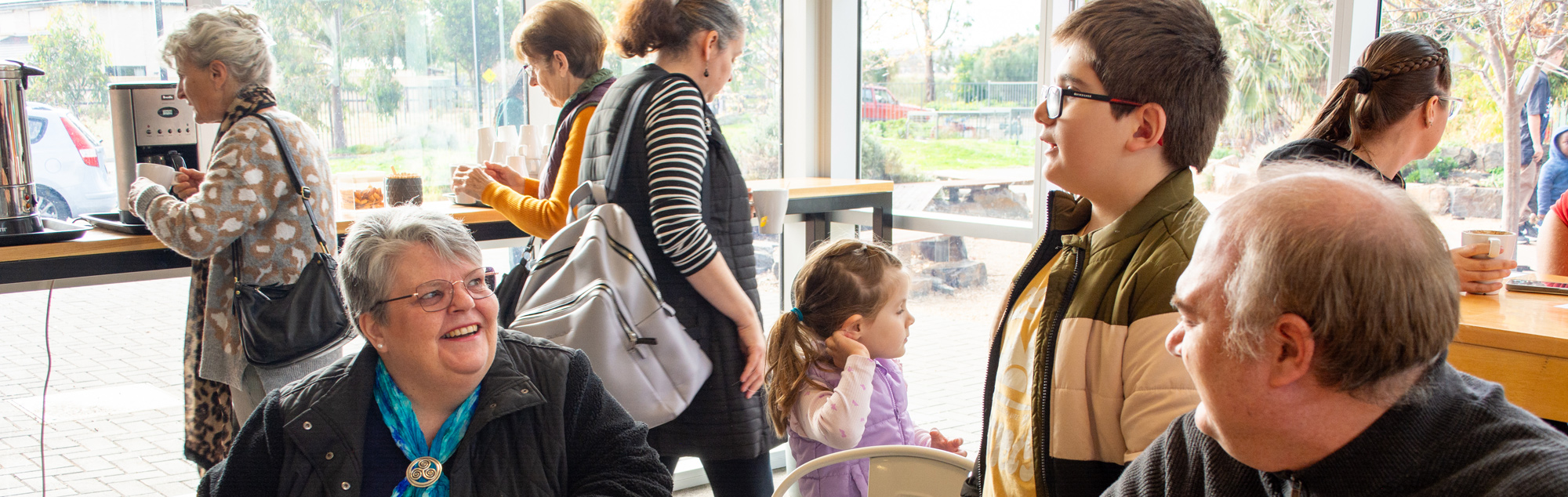 A group of people, both young and old gather together in the foyer. Some engaged in conversation, smiling and laughing together, as coffee is being poured in the background.