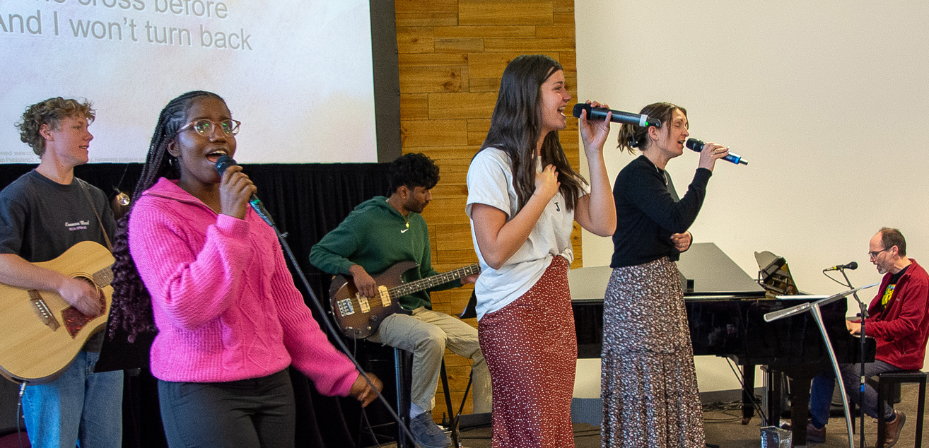 The worship team stands on the stage, microphones and instruments in hand as the sing together praising God.