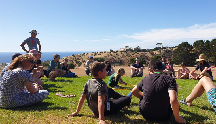 A large group of people, young and old, sitting together outside in the grass enjoying the sun and conversation.