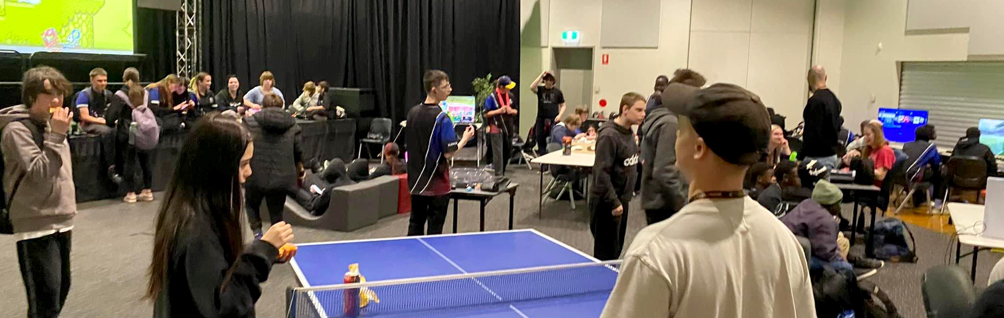 A group of young people gather together in an open space indoors. They are standing together around ping pong tables, and a stage, eating, drinking and chatting.