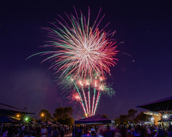 Large fireworks explode in the night sky above a large crowd of people standing together watching in awe.