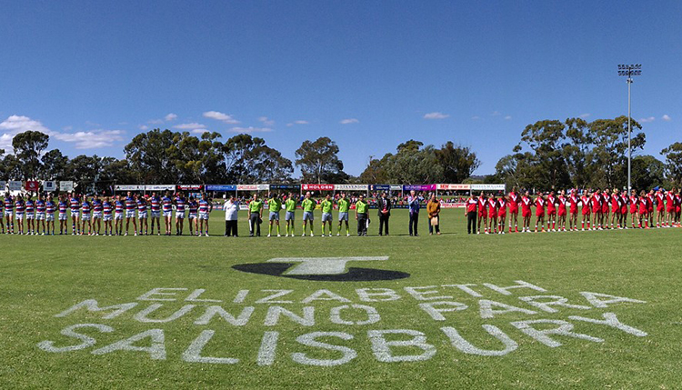 Two full football teams stand lined up in excitement, along with umpires and officials, on a football oval ready for the game to being. 