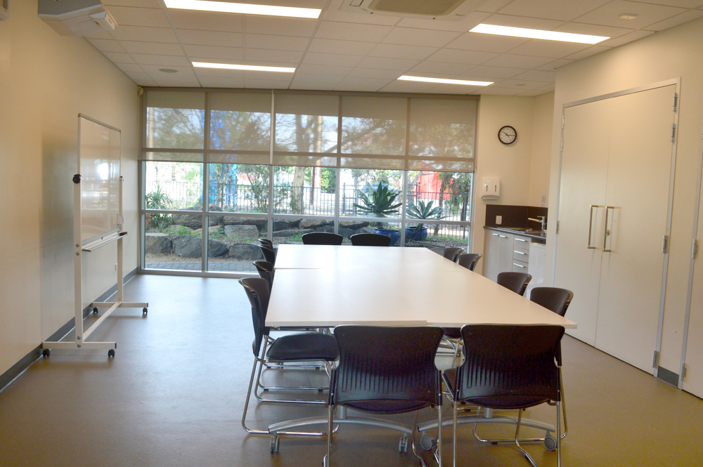 The multipurpose room has a large table in the center of the room, surrounded by chairs. A white board and projector against one wall, with a small kitchenette on the other.  