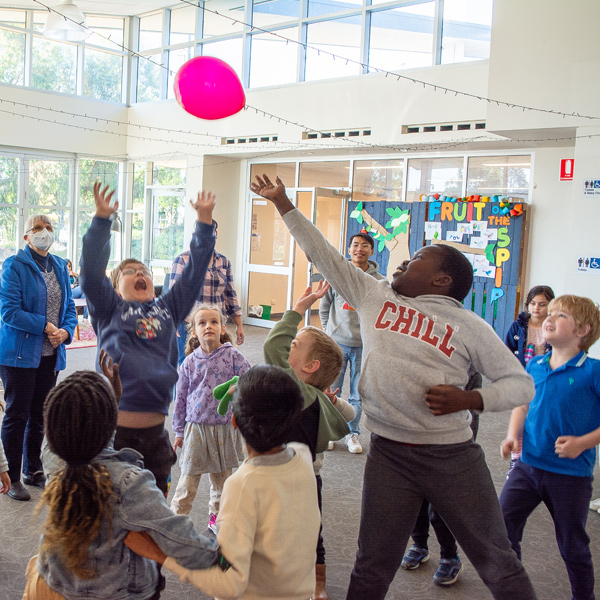 A large group of children are gathered around together playing with a balloon. The ballon is high in the air and the children are jumping and reaching with anticipation and excitement as the balloon slowly falls down within reach.