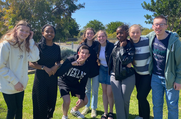 A group of 5 young adult guys standing together with arms around each others shoulders, smiling together.