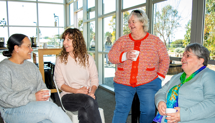 Four women sit together, coffees in hand, smiling, engaged in friendly conversation.