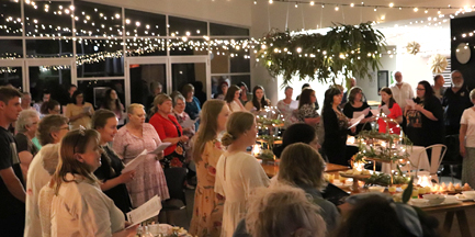 The church foyer is filled with women, standing together, carol books in hand, singing Christmas carols together underneath sparkling fairy lights, surrounded by tables filled with delicious food.